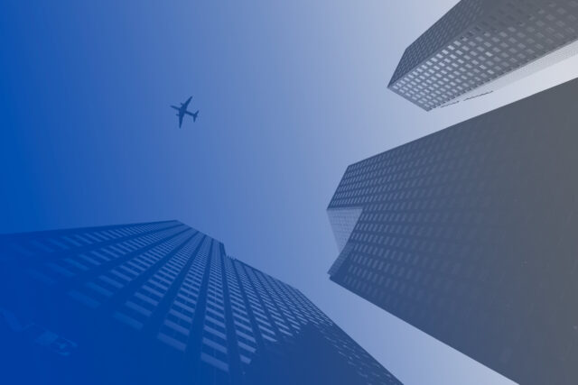 Blue-tinted image looking up at tall skyscrapers with a clear sky in the background, featuring an airplane flying overhead.