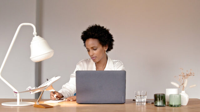 Image of a woman sitting at her wooden desk with decorations, a model airplane, a white lamp and a grey macbook