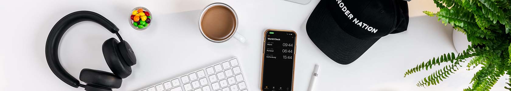 Cropped image of a keyboard, cup of coffee, black headphones, a Noder Nation cap and phone on a white desk