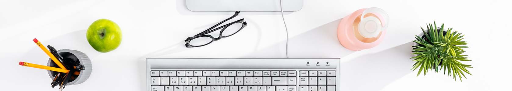 Cropped image of a white desk surface with a green plant and apple, keyboard, pink water bottle, glasses and a pot of pens