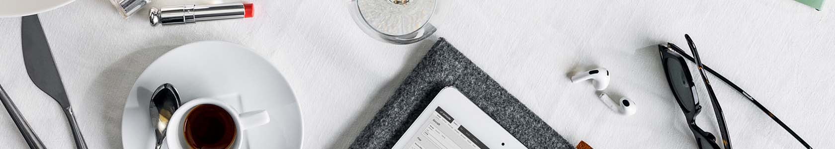 Cropped image of a table with a coffee cup, tablet, glasses, earphones and lipstick on a white table